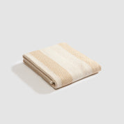 The Herringbone Hues Throw Blanket by In2Green blends sustainability and elegance. Made in the USA from recycled cotton, this Eco throw showcases folded beige and white striped fabric against a neutral backdrop, making it a perfect addition to any space.