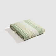 A folded Herringbone Hues Throw Blanket by In2Green, featuring green and white stripes on a plain background, made from recycled cotton.