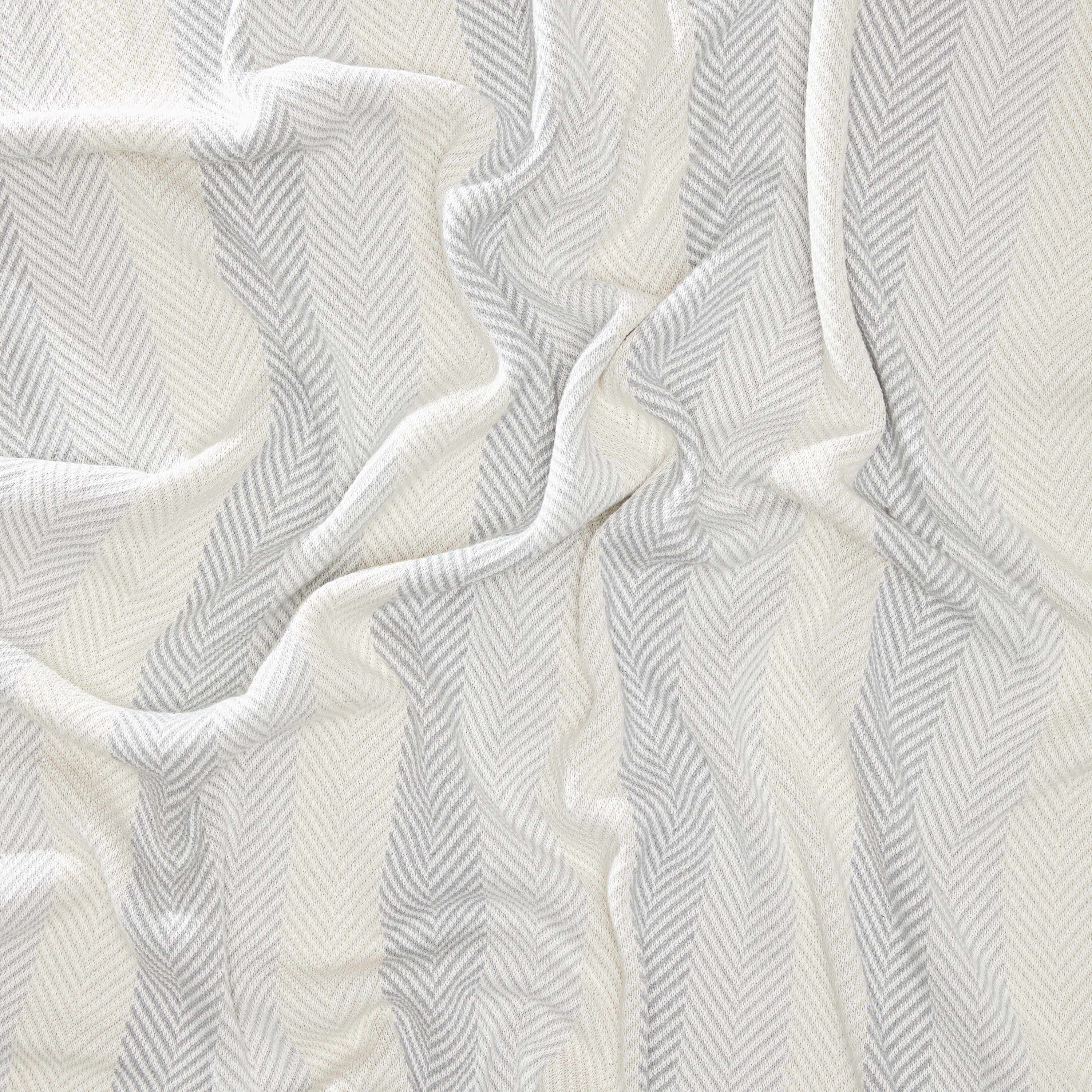 The Herringbone Hues Throw Blanket by In2Green is crafted in the USA from recycled cotton, featuring a wavy drape with a chevron pattern in alternating shades of cream and light gray.