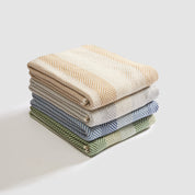 A stack of four In2Green Herringbone Hues Throw Blankets, made from recycled cotton in beige, gray, blue, and green, elegantly rests on a white surface.