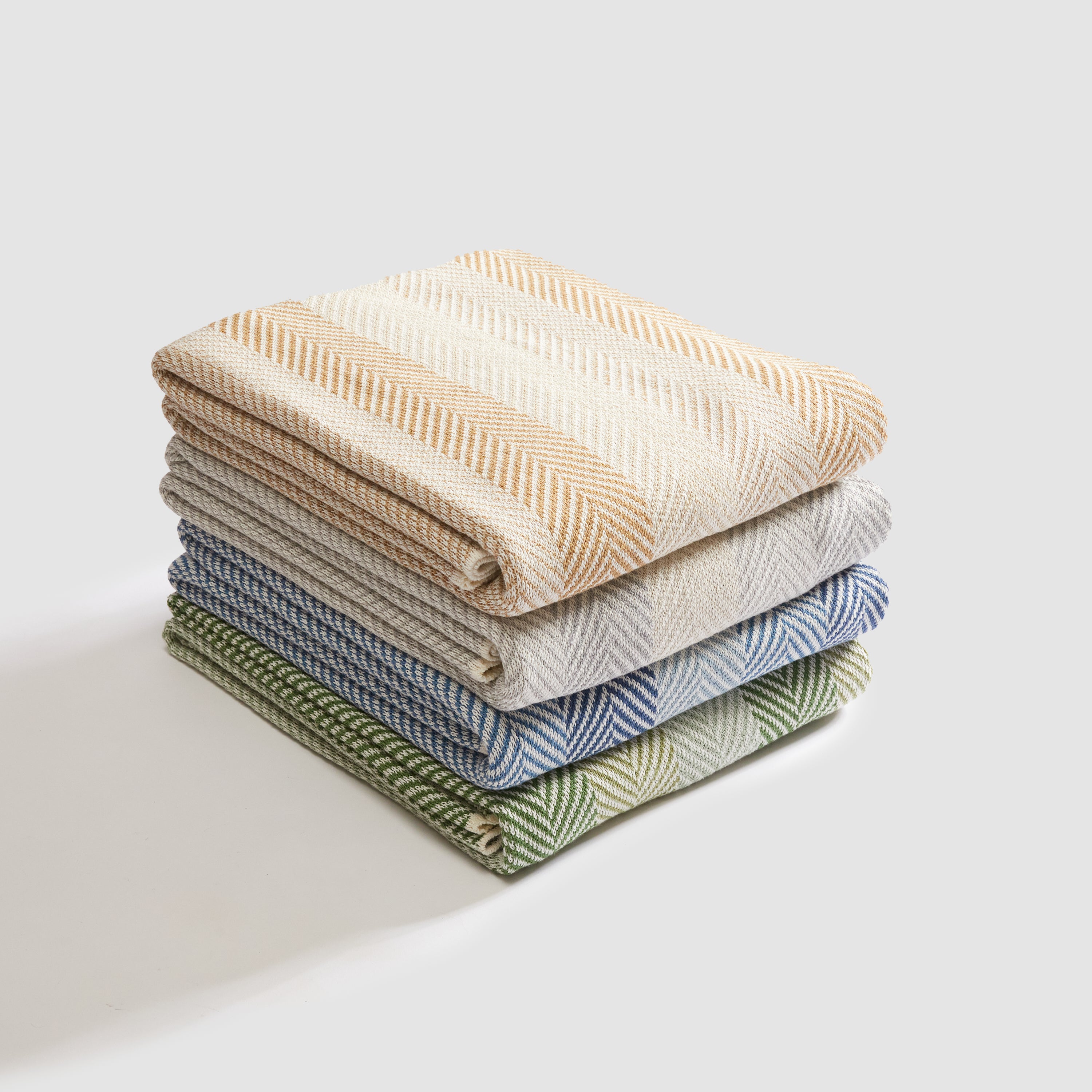 A stack of four In2Green Herringbone Hues Throw Blankets, made from recycled cotton in beige, gray, blue, and green, elegantly rests on a white surface.