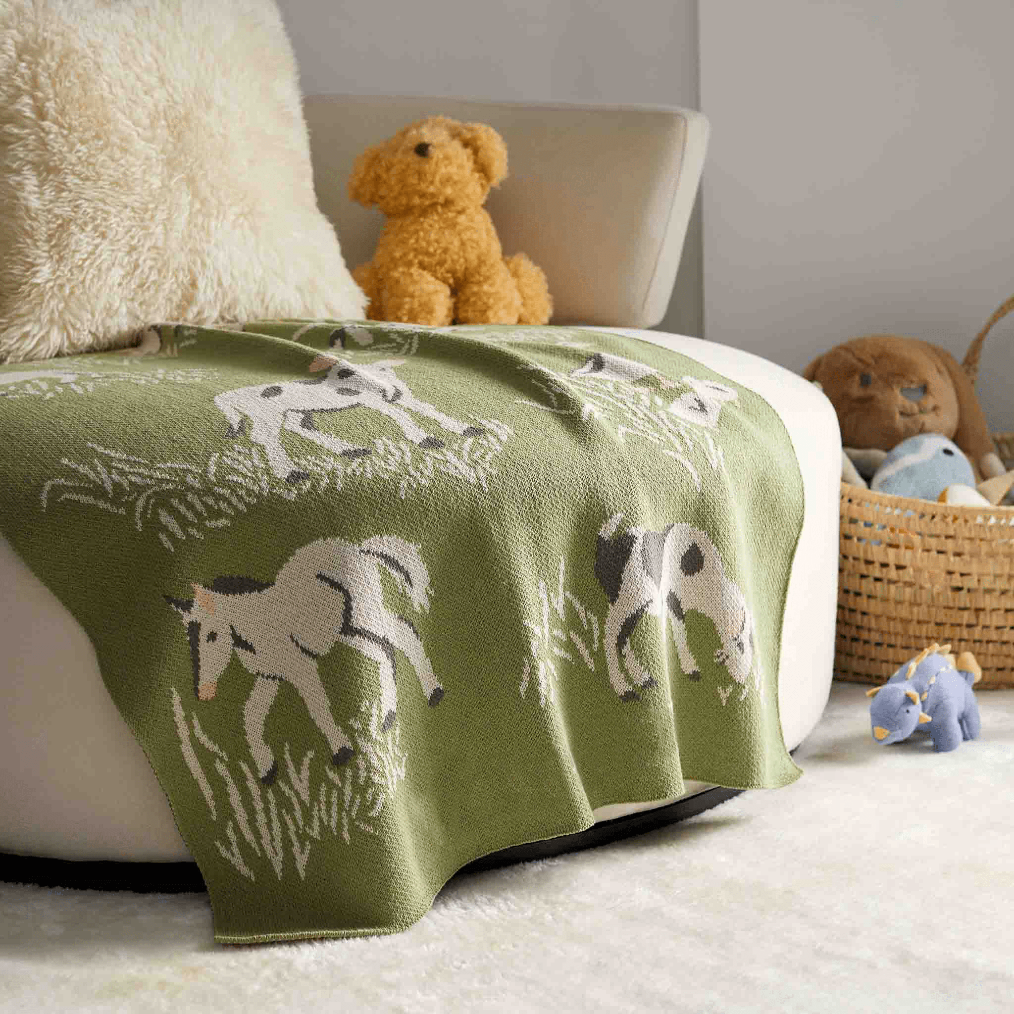 The green in2green Baby Farm Blanket, adorned with horse designs, is draped over a white chair beside a plush dog, basket, and small toys on a soft rug, making it perfect for any baby room.