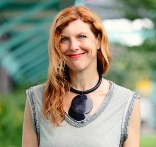 A person with long red hair, dressed in a gray top, accessorized with a striking large black necklace featuring intricate designs, smiles warmly outdoors.