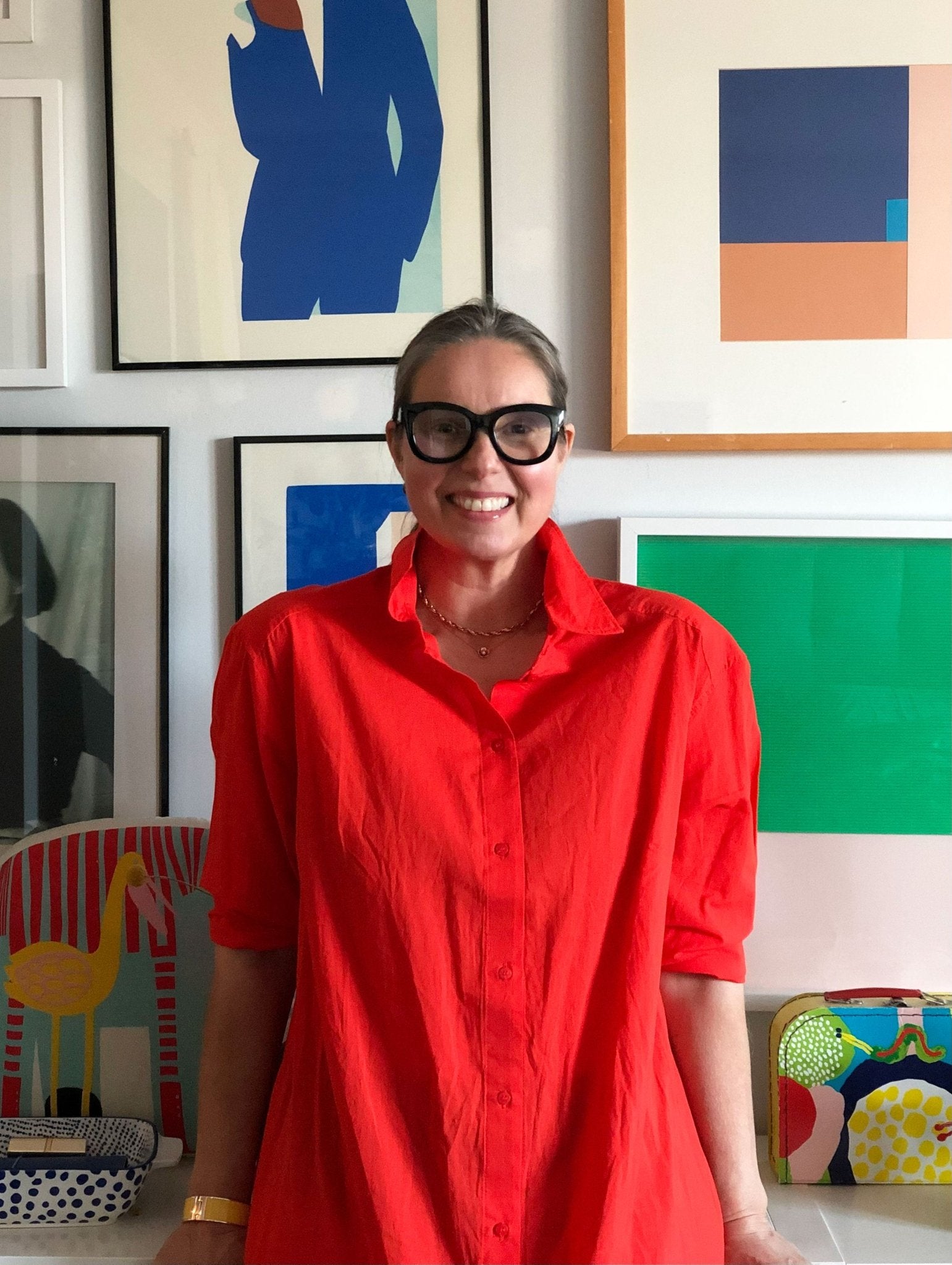 A person in a red shirt and black glasses smiles in front of a wall featuring framed abstract art and the vibrant Poly Kaareva Throw Blanket by Petra Kaksonen from in2green, adding eco-friendly charm.