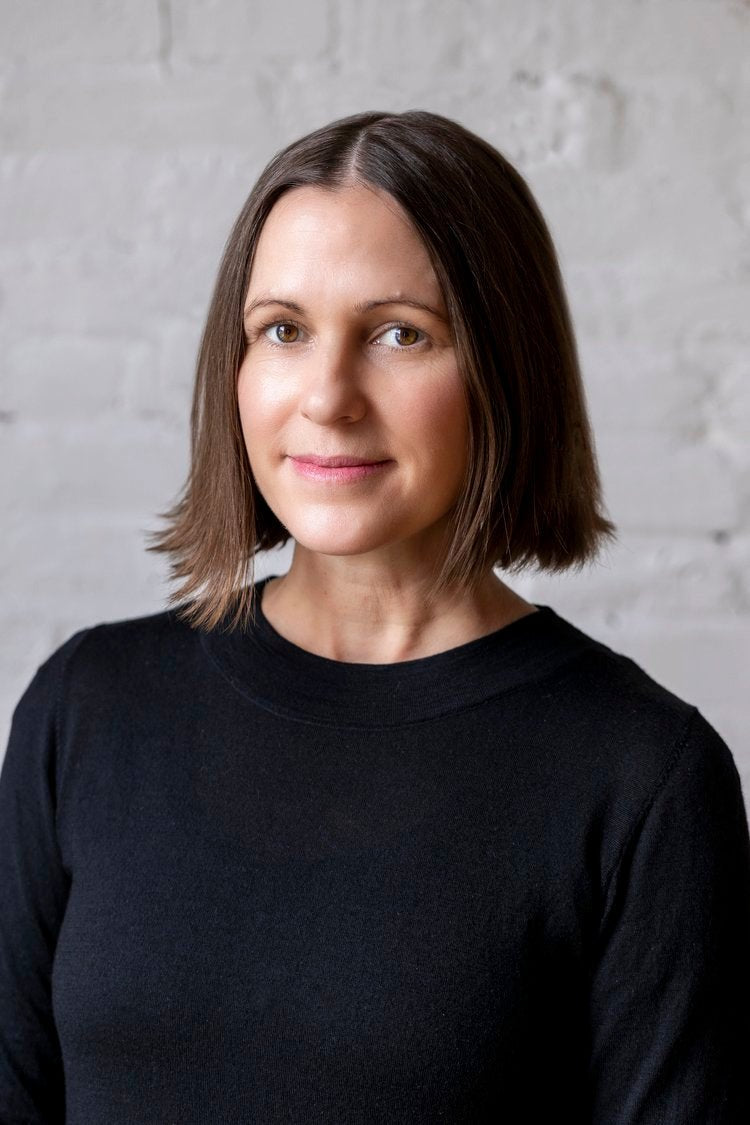 A person with shoulder-length brown hair wearing a black top stands against a white brick wall, reminiscent of an urban landscape, similar to the modern aesthetic of the Level Throw Blanket by Kelly Harris Smith from in2green.