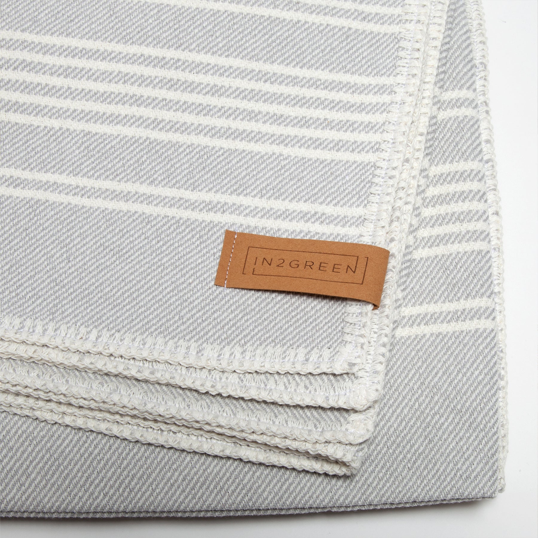 This Woven Twill Blanket, made from recycled cotton and eco yarns, showcases a gray and white striped design with a brown "in2green" label on the corner.